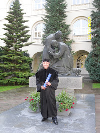 Uśmiechnięty mężczyzna w todze, trzymający dyplom, stoi na tle pomnika i budynku uniwersyteckiego. Widoczne drzewa iglaste i klomby z czerwonymi kwiatami.