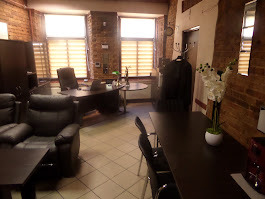 Wnętrze biura z ceglanymi ścianami, ciemnymi meblami. W tle biurko z laptopem, krzesło obrotowe. W rogu wieszak z ubraniami. Uwagę przykuwają białe kwiaty w wazonie.