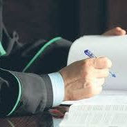 Prawnik w todze podpisuje dokument. Ujęcie z bliska na dłoń z długopisem nad stosem białych kartek i fragment togi prawniczej.