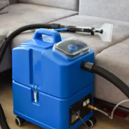 Niebieski odkurzacz piorący z przezroczystą nakładką na zbiorniku, wąż i ssawka na tle szarej, narożnej kanapy. Sprzęt do czyszczenia tapicerki.