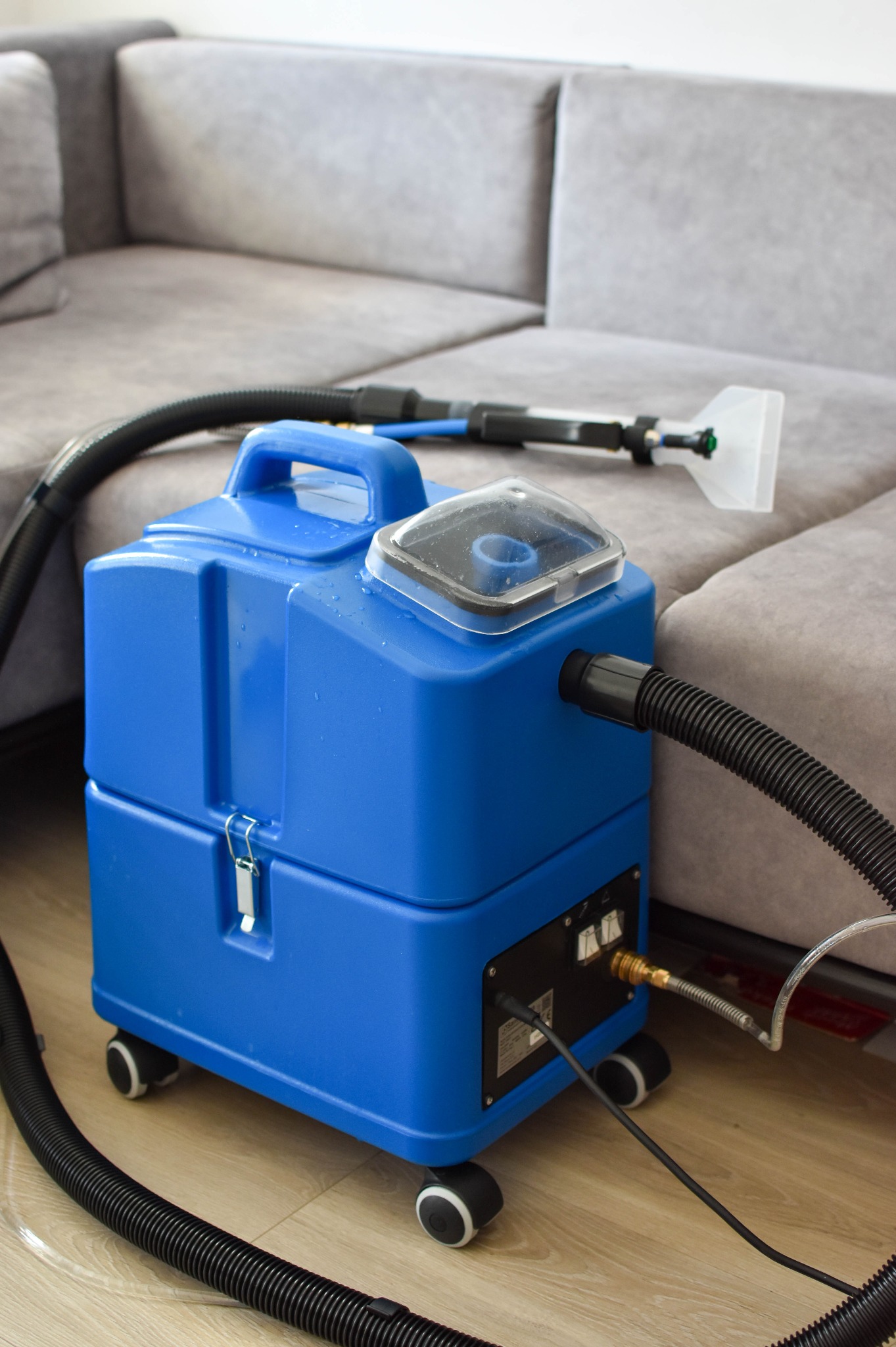 Niebieski odkurzacz piorący z przezroczystą nakładką na zbiorniku, wąż i ssawka na tle szarej, narożnej kanapy. Sprzęt do czyszczenia tapicerki.