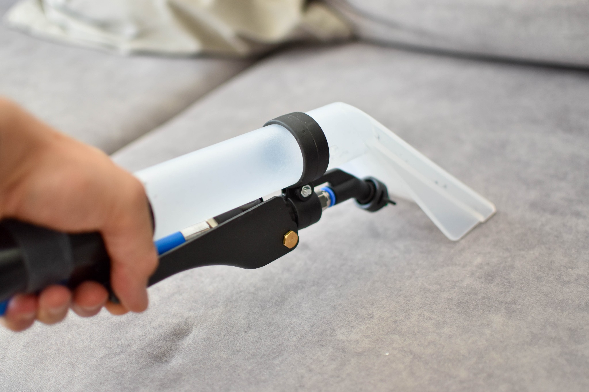 Dysza ekstraktora w dłoni, czyszcząca szarą kanapę. Widoczny fragment sofy i narzędzie do ekstrakcyjnego prania tapicerki.