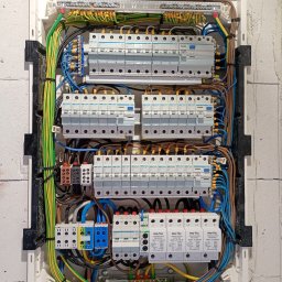 Piotr - Rozdzielnica elektryczna z zabezpieczeniami przepięciowymi Pro-Tec, widoczne przewody w różnych kolorach, zamontowana na ścianie z bloczków.
