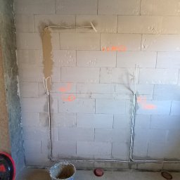Piotr - Ściana z betonu komórkowego z poprowadzonymi przewodami elektrycznymi, oznaczonymi pomarańczowymi napisami. Widoczny fragment podłogi z wiadrem i elementami instalacji.