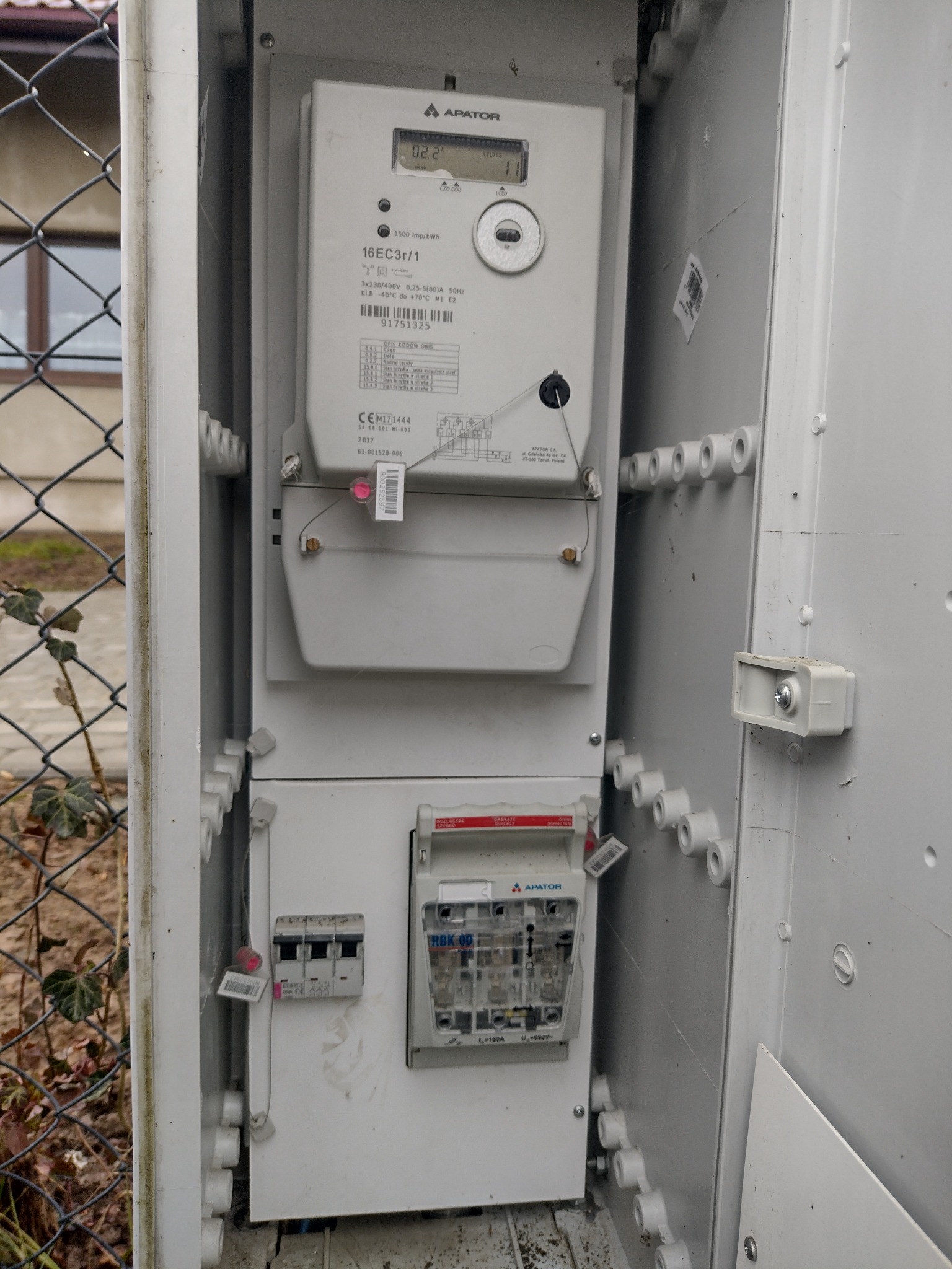 Otwarta skrzynka z licznikiem i bezpiecznikami Apator, widoczne plombowanie. Element instalacji elektrycznej na tle siatki ogrodzeniowej i zieleni.