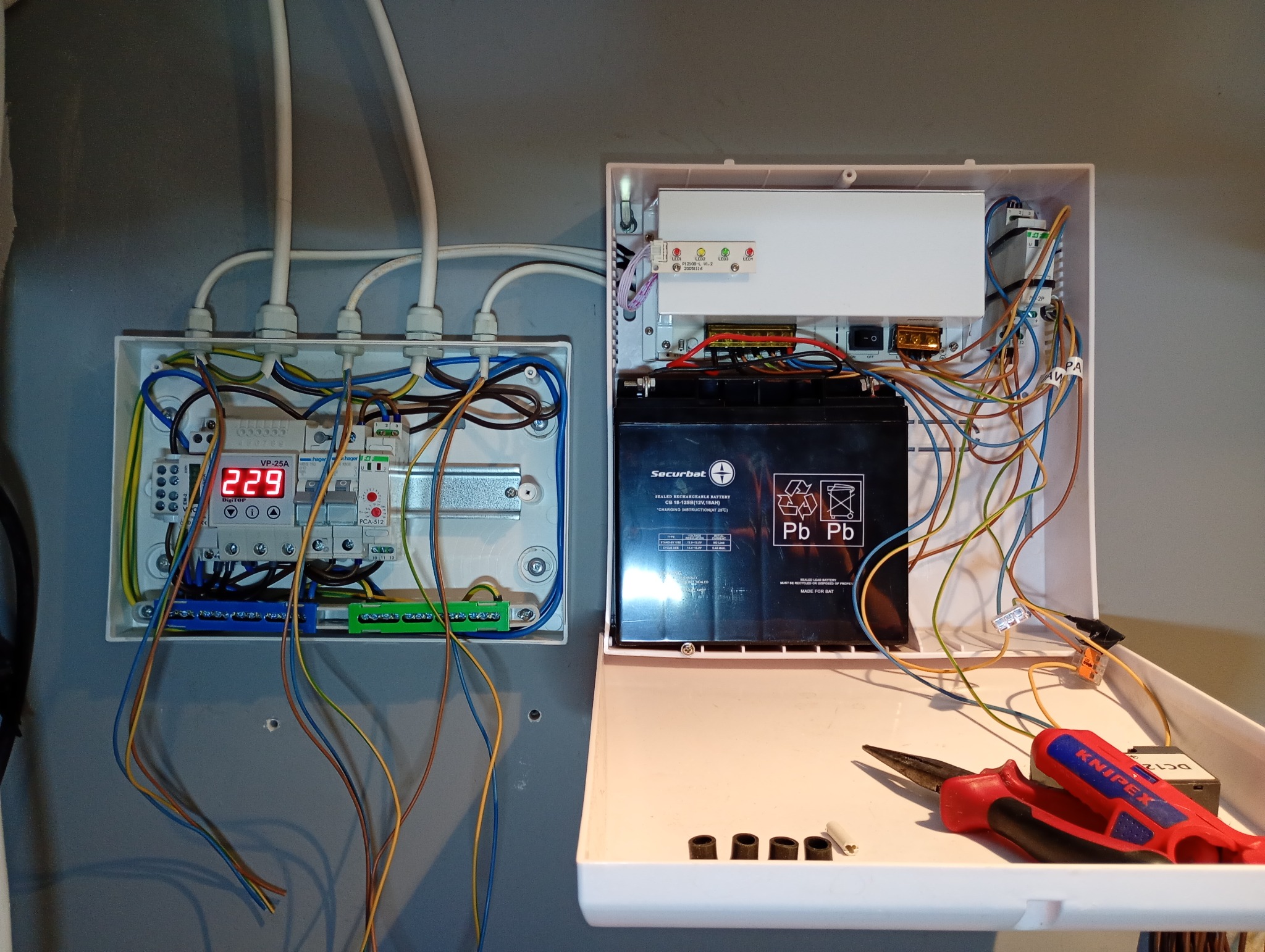 Otwarta skrzynka z akumulatorem i elektroniką alarmową, obok druga skrzynka z licznikiem energii elektrycznej i przewodami. Widoczne narzędzia.