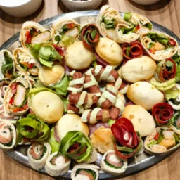 Srebrna patera z roladkami tortilli z łososiem, kurczakiem i warzywami, mini kanapeczkami i kiełbaskami w cieście francuskim, na drewnianym stole.