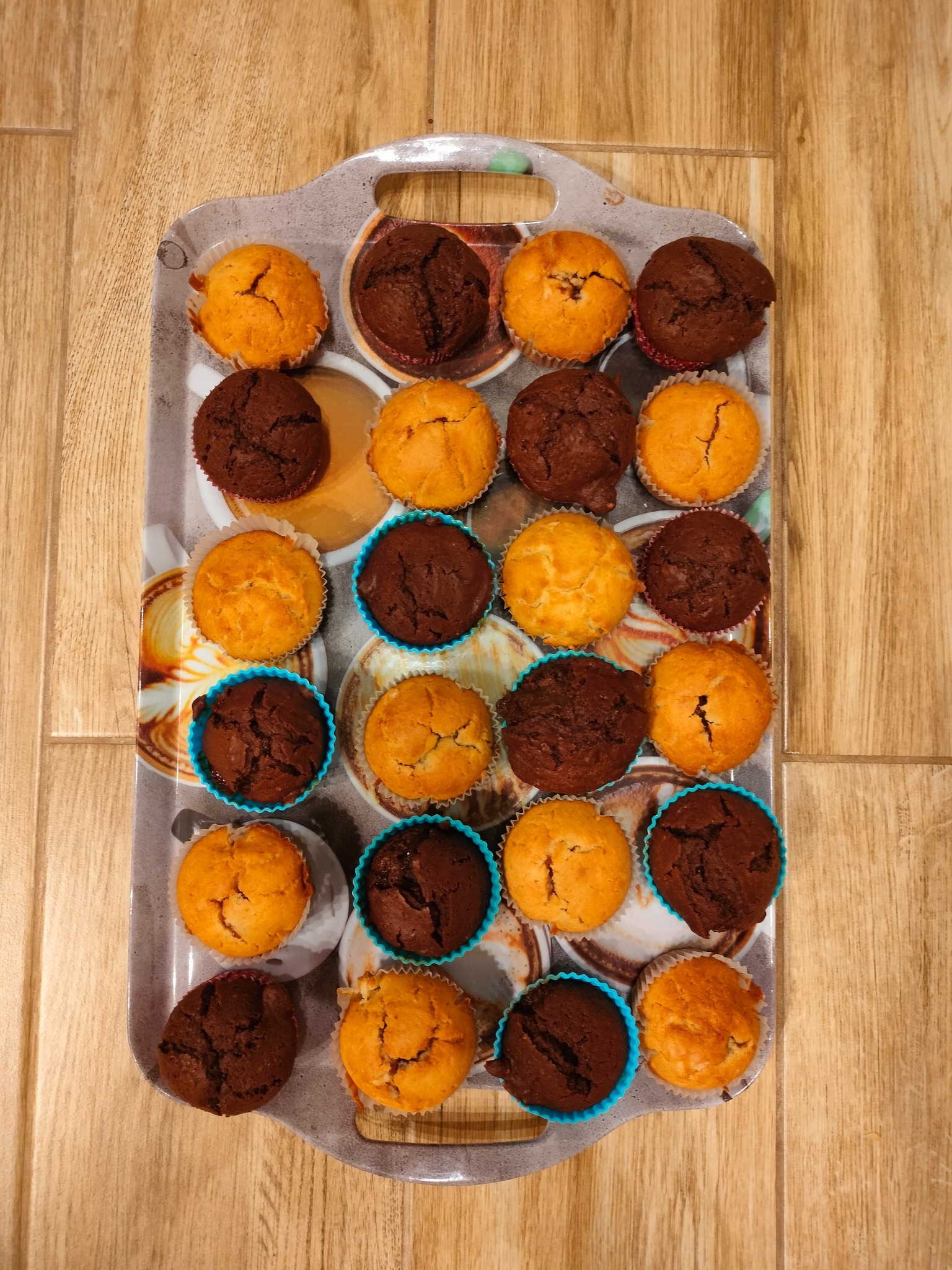 Taca z babeczkami: jasne i czekoladowe muffiny w papilotkach, ułożone rzędami na tacy z motywem kawy, na drewnianym tle. Widok z góry, ciepłe światło.