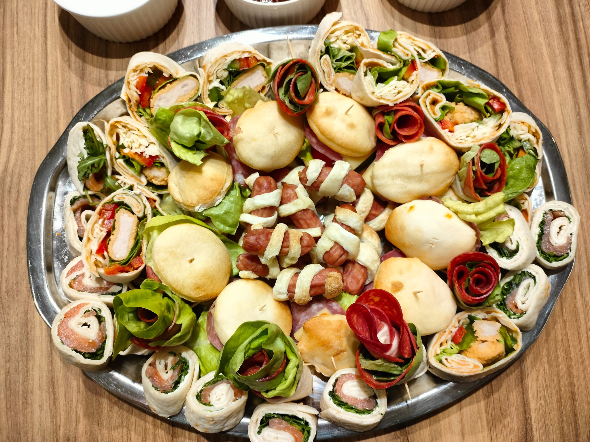 Srebrna patera z roladkami tortilli z łososiem, kurczakiem i warzywami, mini kanapeczkami i kiełbaskami w cieście francuskim, na drewnianym stole.