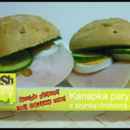 Dwie kanapki paryskie z szynką drobiową, jajkiem i og&oacute;rkiem, pieczywo z widocznymi nasionami, na ż&oacute;łtym tle z napisem reklamowym.