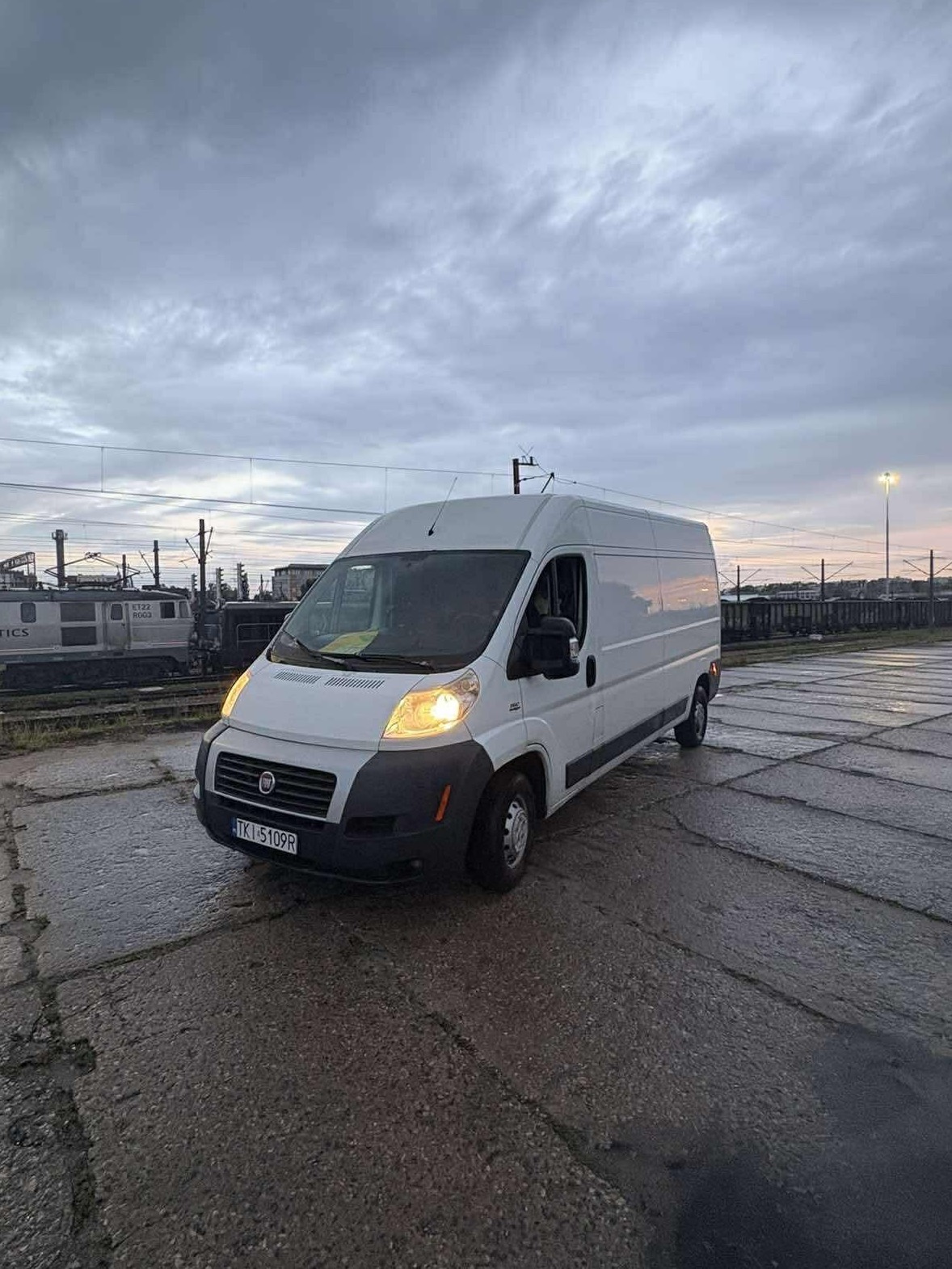 Biały furgon Fiat Ducato na betonowym placu, światła włączone, w tle tory kolejowe i pochmurne niebo, widok z przodu pod lekkim kątem.
