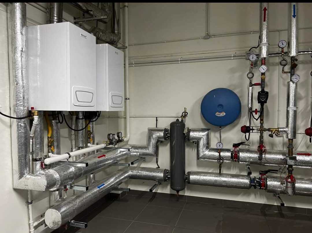 Dwa białe piece CO wiszące nad rzędami rur z izolacją termiczną, manometry i zawory. Instalacja grzewcza w kotłowni z szarą podłogą.