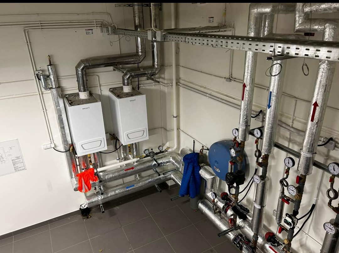 Dwa białe piece CO zamontowane w kotłowni, z widocznymi rurami, manometrami i osprzętem. Na ścianie widoczny schemat instalacji. Czerwone i niebieskie akcenty na rurach.