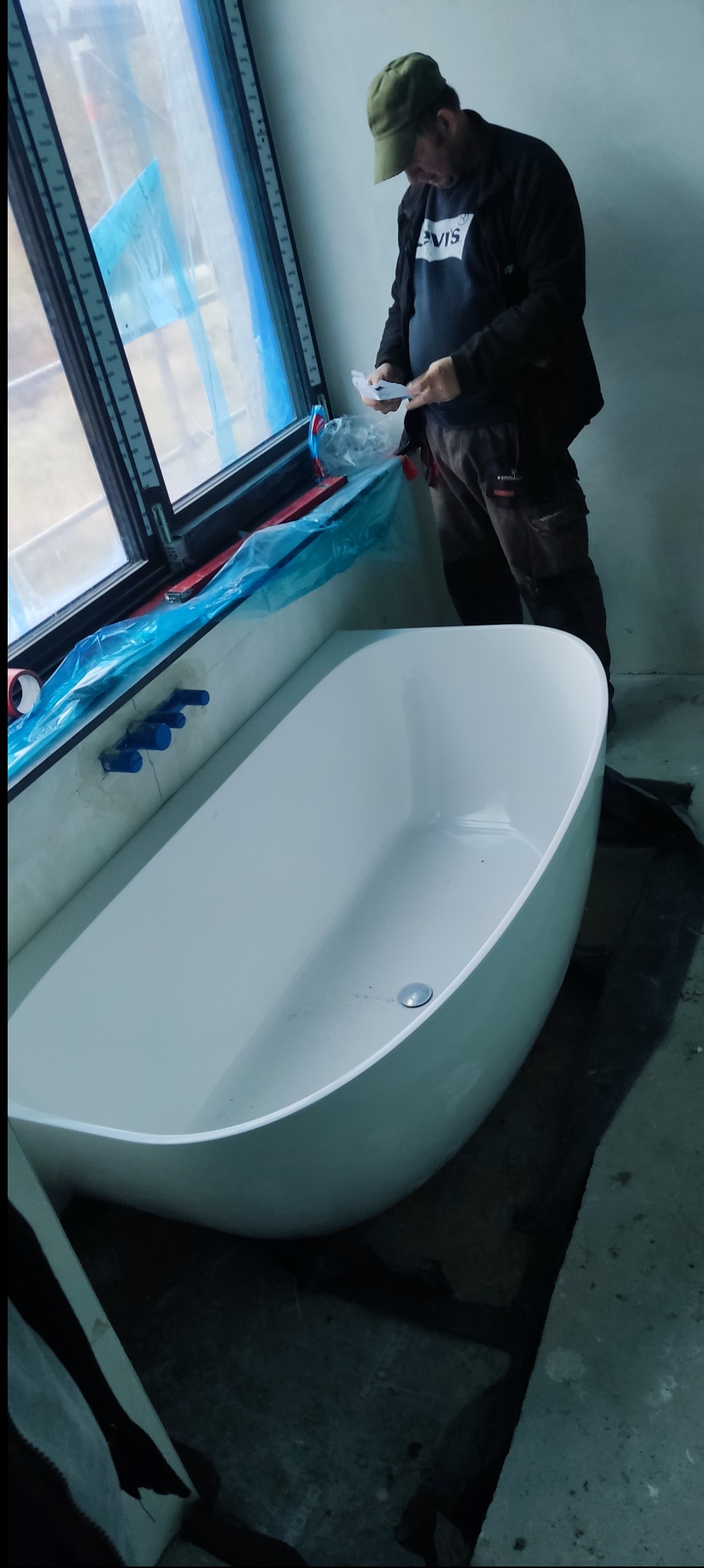 Biała wanna wolnostojąca w łazience w trakcie remontu, przy oknie zabezpieczonym folią. Mężczyzna w tle rozpakowuje elementy montażowe.