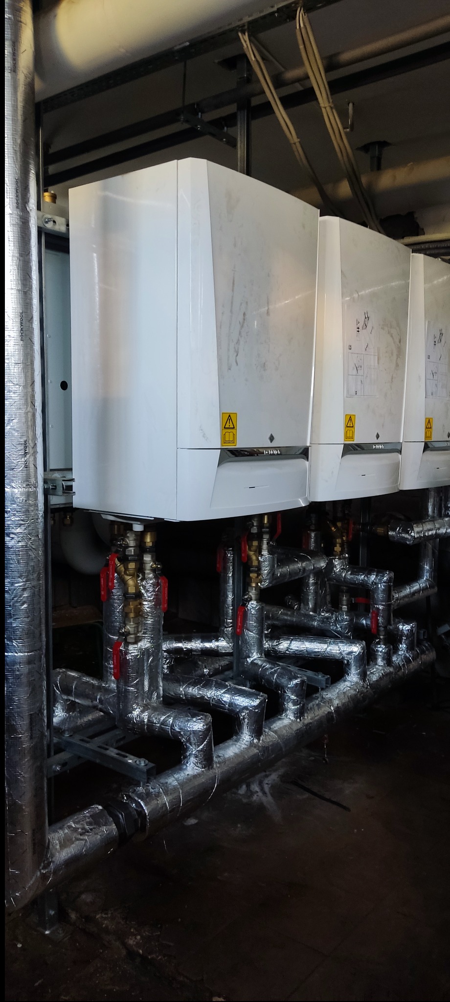 Rząd białych, nowoczesnych pieców CO z izolowanymi rurami i zaworami z czerwonymi uchwytami. Widok na instalację grzewczą w kotłowni.