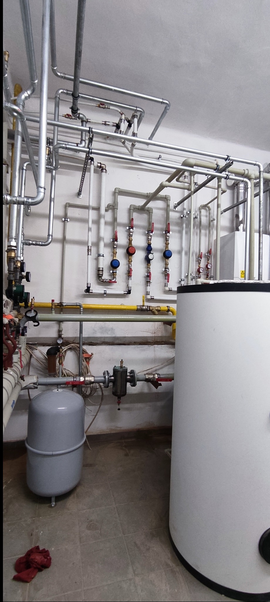 Złożona instalacja CO z rur stalowych, zaworów i zbiornika wyrównawczego w kotłowni. Widoczne liczne połączenia i elementy armatury. Ujęcie z dołu, ukazujące szczegóły instalacji.