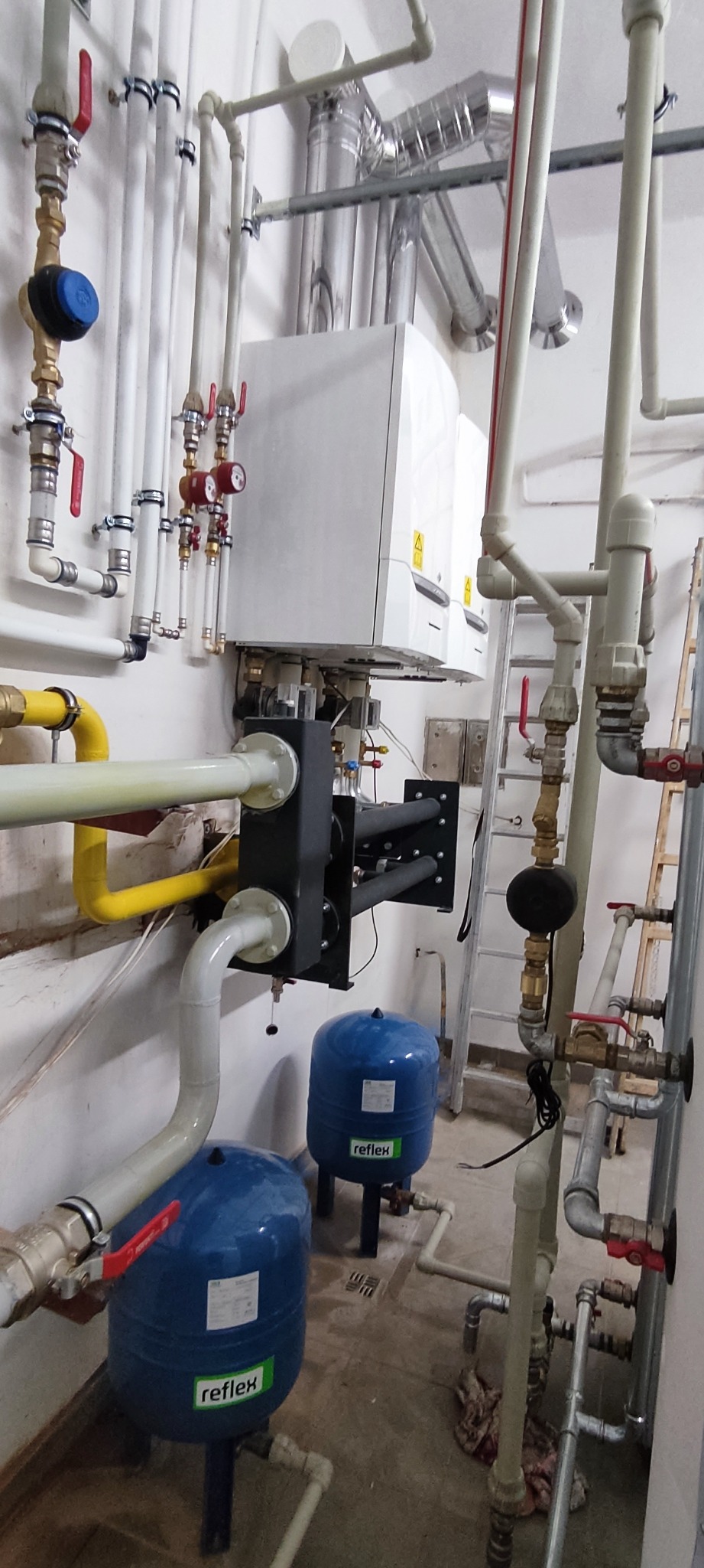 Biały piec CO z licznymi rurami, zaworami i dwoma niebieskimi zbiornikami Reflex na tle białej ściany. Widoczna drabina. Instalacja po montażu, gotowa do użytku.