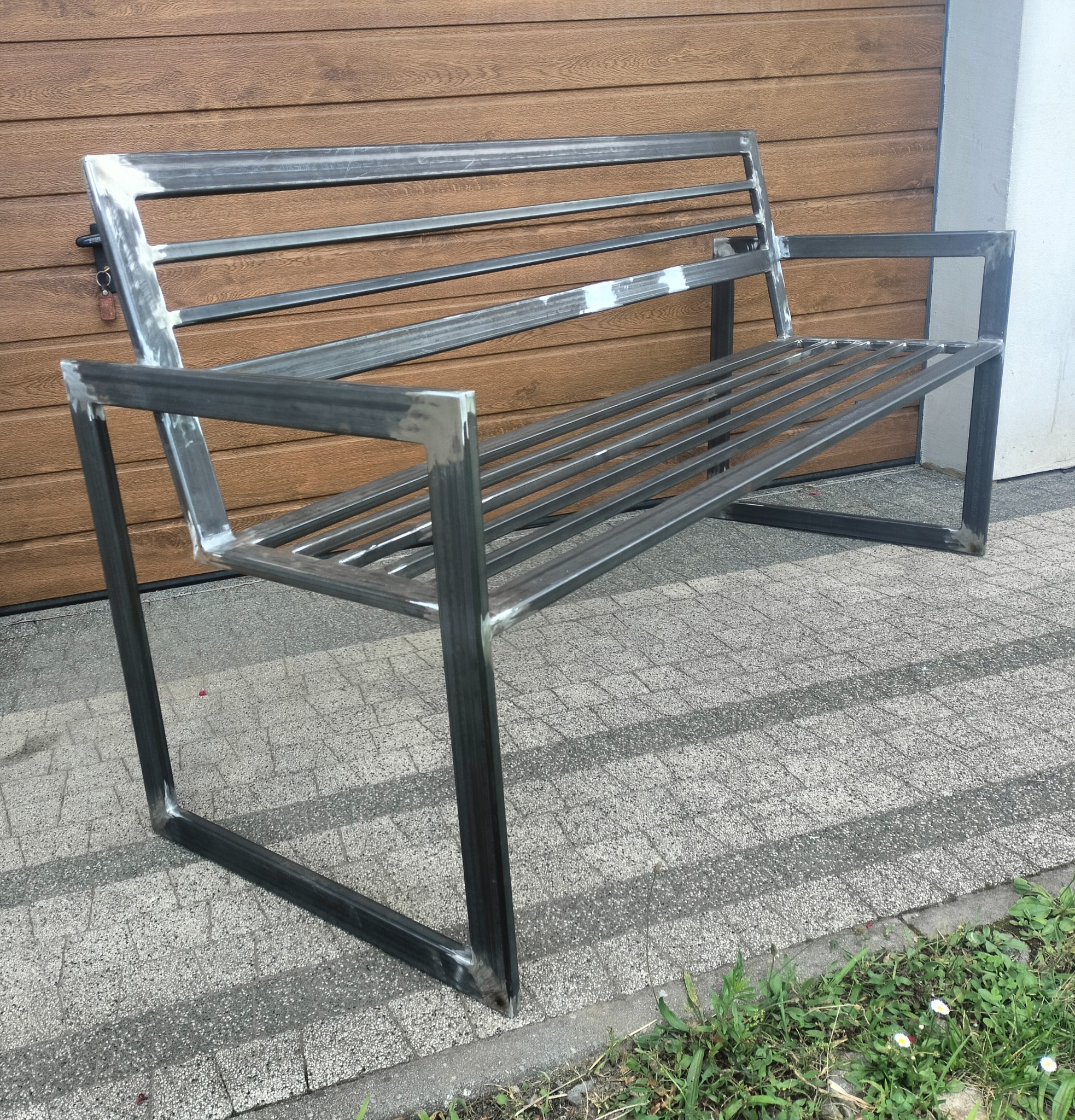 Metalowa ławka ogrodowa z profili, spawana, o surowym wykończeniu, na tle drewnianej bramy garażowej i kostki brukowej. Widoczne ślady po spawaniu.