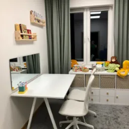 Gabinet logopedyczny w Olsztynie: białe biurko z lustrem, krzesła na kółkach, półki z książkami i zabawki na szafce przy oknie. Przyjazna przestrzeń.