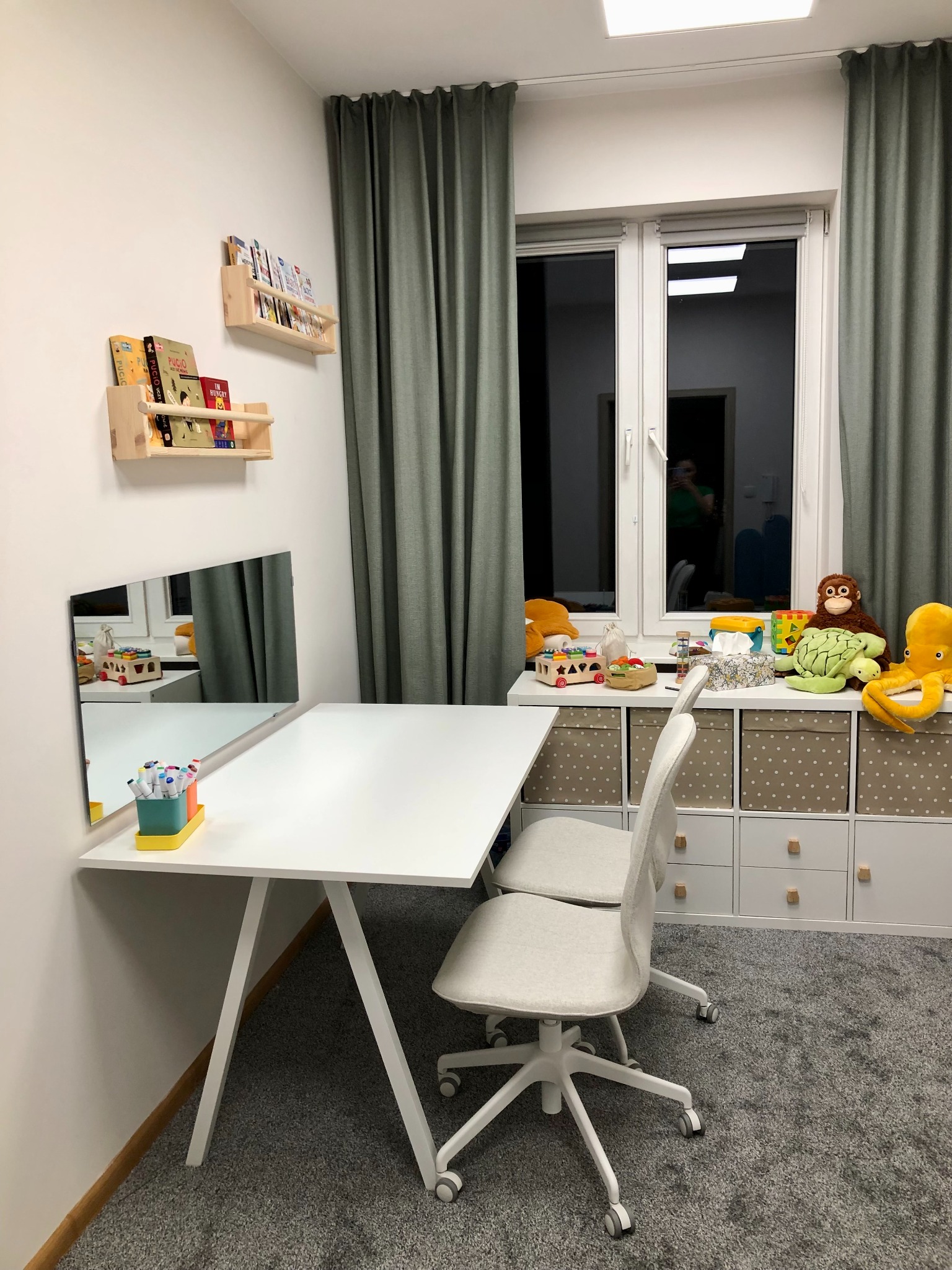 Gabinet logopedyczny w Olsztynie: białe biurko z lustrem, krzesła na kółkach, półki z książkami i zabawki na szafce przy oknie. Przyjazna przestrzeń.