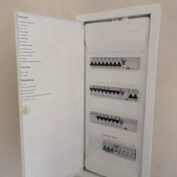 Rozdzielnica elektryczna w białej obudowie z otwartymi drzwiczkami, na których widoczna jest lista obwodów. Widoczne bezpieczniki i wyłącznik główny.