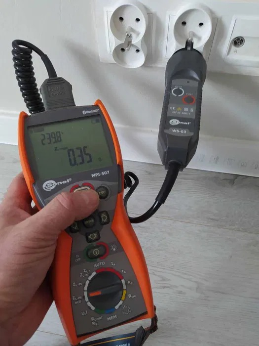 Elektryk wykonuje pomiary elektryczne w gniazdkach za pomocą miernika marki Sonel MPI-507 i testera WS-03. Widoczne wartości napięcia i rezystancji na wyświetlaczu.