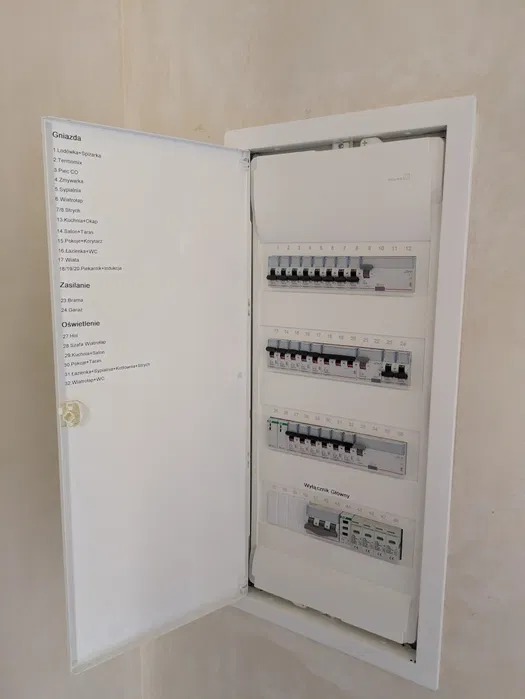 Rozdzielnica elektryczna w białej obudowie z otwartymi drzwiczkami, na których widoczna jest lista obwodów. Widoczne bezpieczniki i wyłącznik główny.