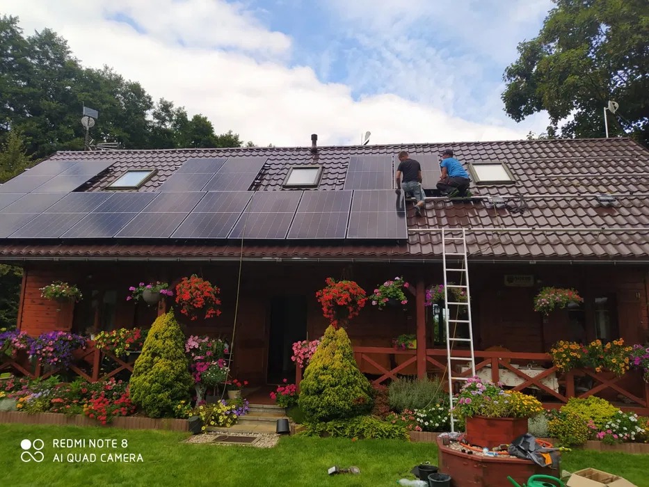 Montaż paneli słonecznych na dachu drewnianego domu z kwiatami. Dwóch monterów na drabinie instaluje panele fotowoltaiczne. Widok na dach i fragment ogrodu.