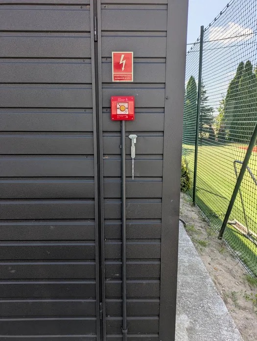 Czerwony przycisk awaryjny odłączający zasilanie, umieszczony na ciemnej, poziomo żebrowanej ścianie budynku. Nad nim znak ostrzegawczy z symbolem błyskawicy.