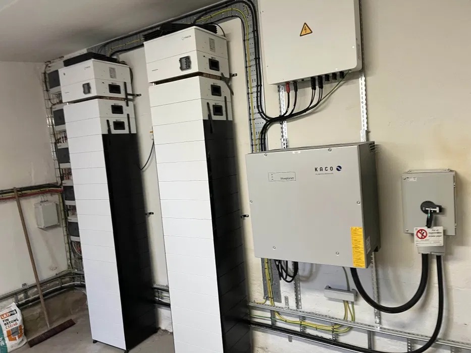 Zestaw akumulatorów i falowników Kaco na ścianie w pomieszczeniu technicznym. Widoczne okablowanie i oznakowania ostrzegawcze. Instalacja fotowoltaiczna.