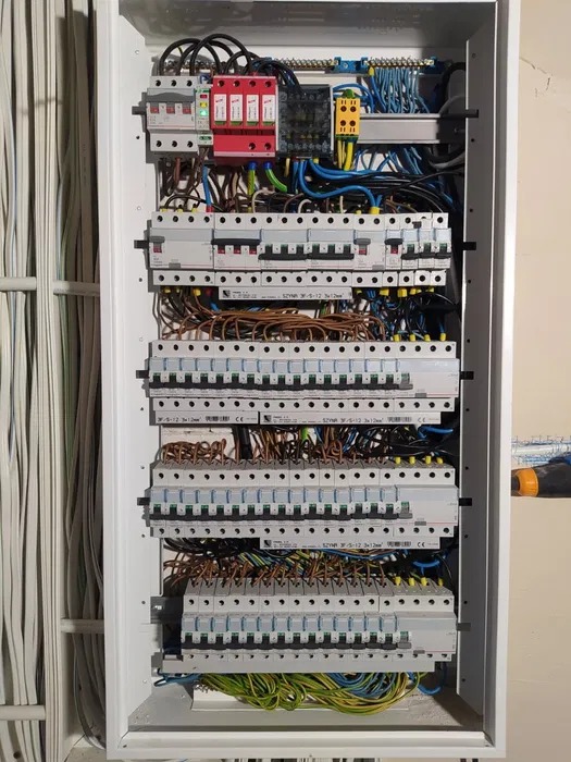 Rozdzielnica elektryczna w białej obudowie, z widocznymi bezpiecznikami, przewodami i elementami zabezpieczeń. Widoczne złącza i kable.