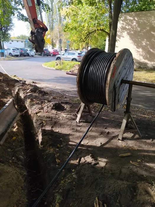 Wykop z kablem na drewnianej szpuli, prawdopodobnie w trakcie układania, z fragmentem koparki w tle i widokiem na ulicę.
