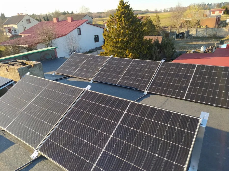 Panele fotowoltaiczne zamontowane na płaskim dachu budynku mieszkalnego z widocznym kominem i otoczeniem wiejskim w tle.