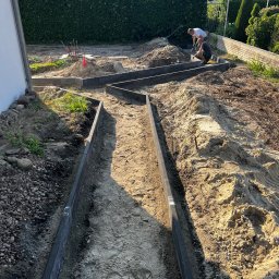 RAFAŁ KAMIŃSKI - Prace ziemne przy zakładaniu ogrodu: dwóch mężczyzn montuje obrzeża rabat w zygzakowatym układzie. Widoczne narzędzia i przygotowany teren pod nasadzenia.