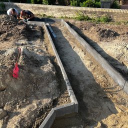 RAFAŁ KAMIŃSKI - Montaż obrzeży trawnikowych z szarego betonu przez dwóch mężczyzn. W tle zielona roślinność i ceglany murek. Widoczne narzędzia i poziomica.