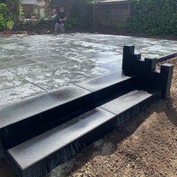 RAFAŁ KAMIŃSKI - Układanie mokrych, szarych płyt chodnikowych na zewnątrz. W tle mężczyzna wyrównuje podłoże pod kolejne płyty. Widoczne czarne obrzeża.