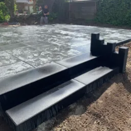 Układanie mokrych, szarych płyt chodnikowych na zewnątrz. W tle mężczyzna wyrównuje podłoże pod kolejne płyty. Widoczne czarne obrzeża.