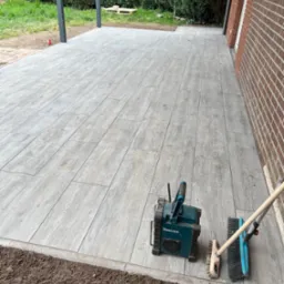 Nowo ułożona, drewnopodobna, szara kostka brukowa na tarasie, z widocznymi narzędziami pracy (szczotki, radio) i świeżo przygotowanym obrzeżem pod nasadzenia.