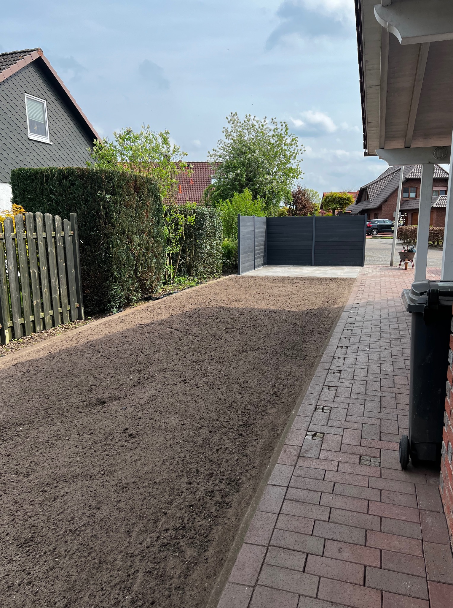 Przygotowany teren pod trawnik z widoczną świeżą ziemią, obok ścieżka z kostki brukowej i czarny śmietnik. W tle ogrodzenie i budynki mieszkalne.