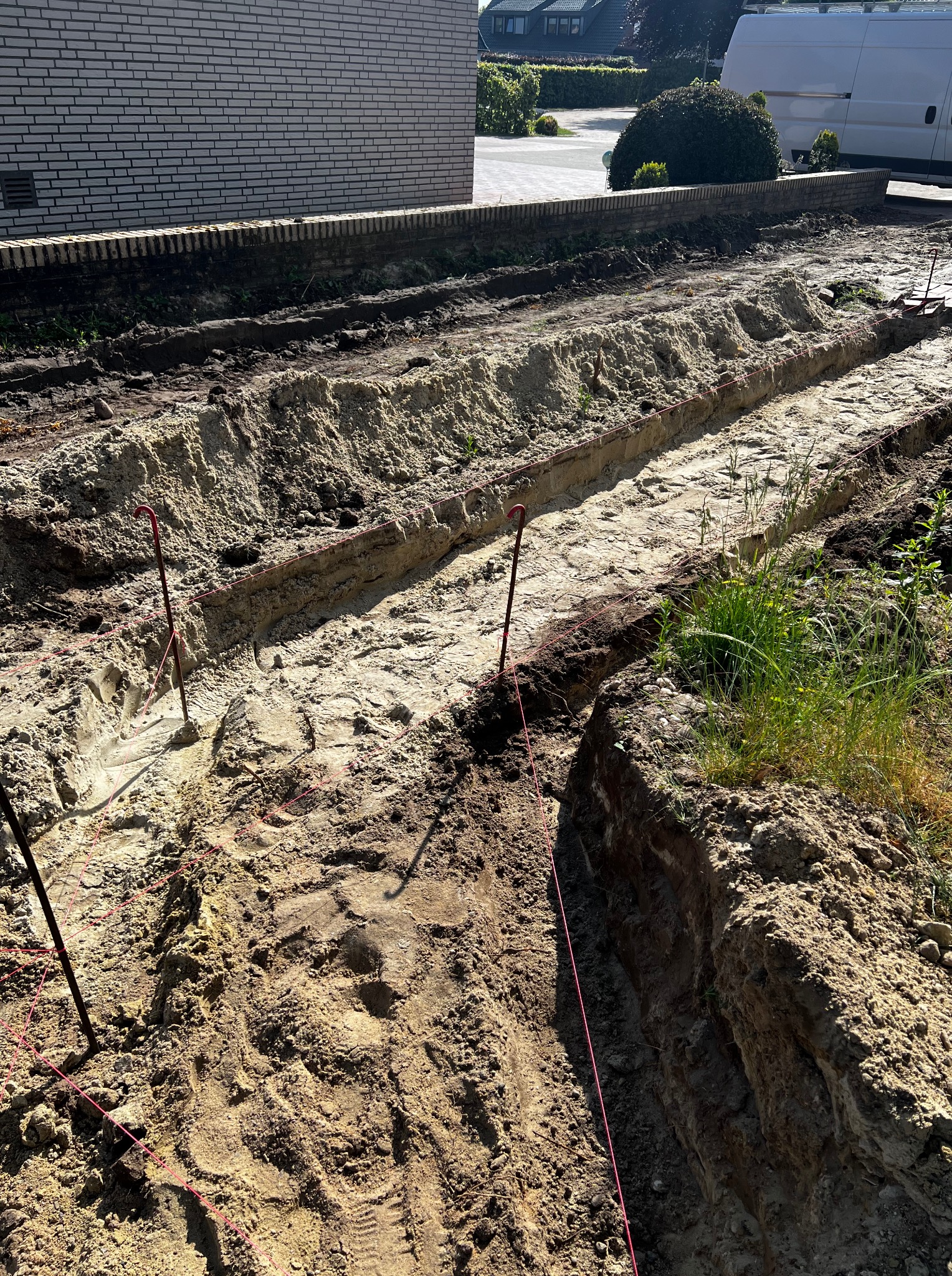 Przygotowanie terenu pod ogród: wykopane rowy z wyznaczonymi granicami czerwoną linką, widoczne ślady opon, element muru w tle i budynki w oddali.