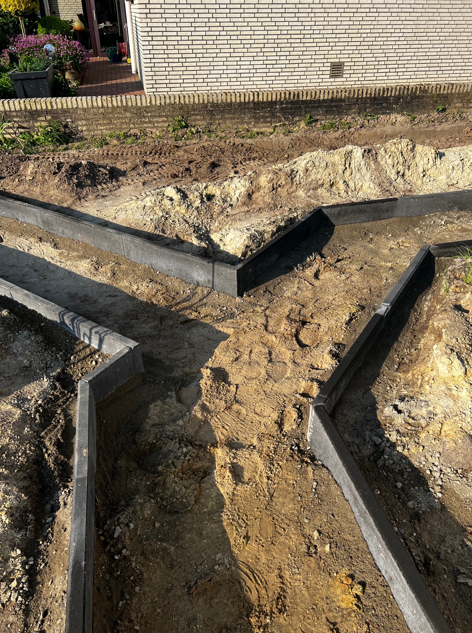 Etap tworzenia ścieżki ogrodowej z szarych obrzeży, widok z góry na wykopaną ziemię i fragment budynku w tle, oświetlone słońcem.