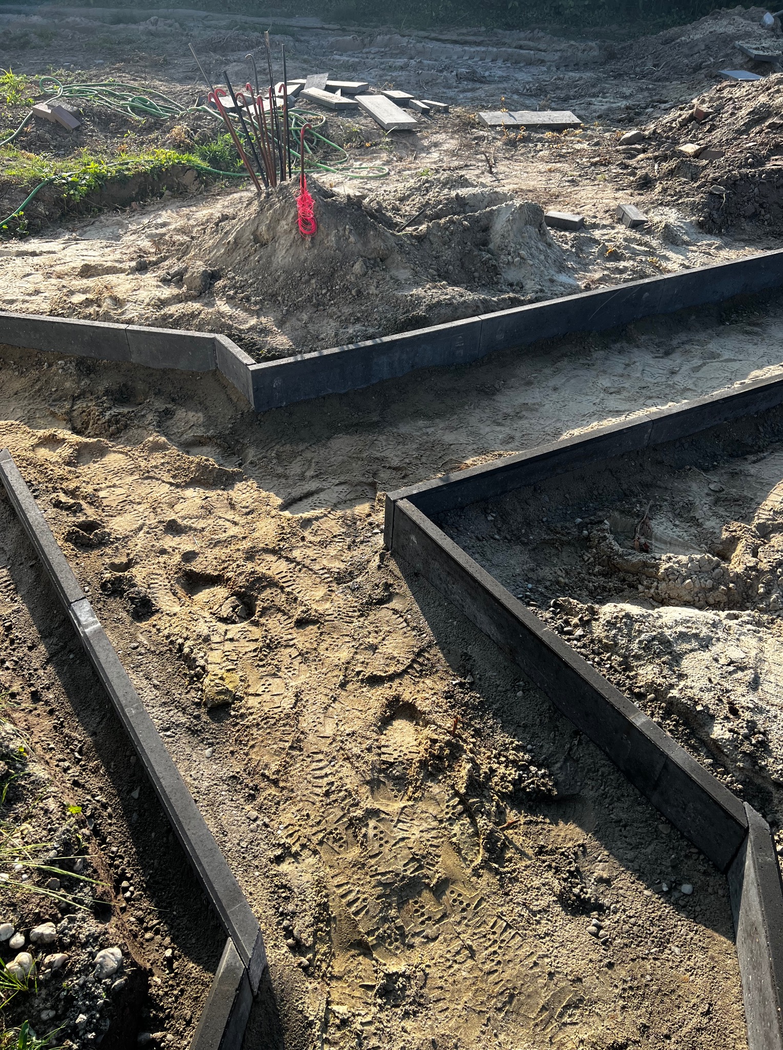 Etap budowy ścieżki ogrodowej z wykorzystaniem obrzeży z kostki. Widoczne ślady stóp na piasku, przygotowane podłoże i narzędzia.