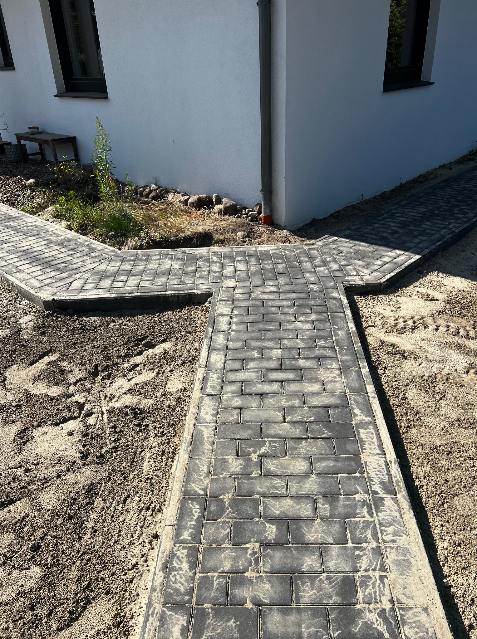 Świeżo ułożona, szara kostka brukowa tworząca ścieżkę rozwidlającą się na trzy strony, z widocznymi śladami pracy w postaci rozsypanego piasku i fragmentu budynku w tle.