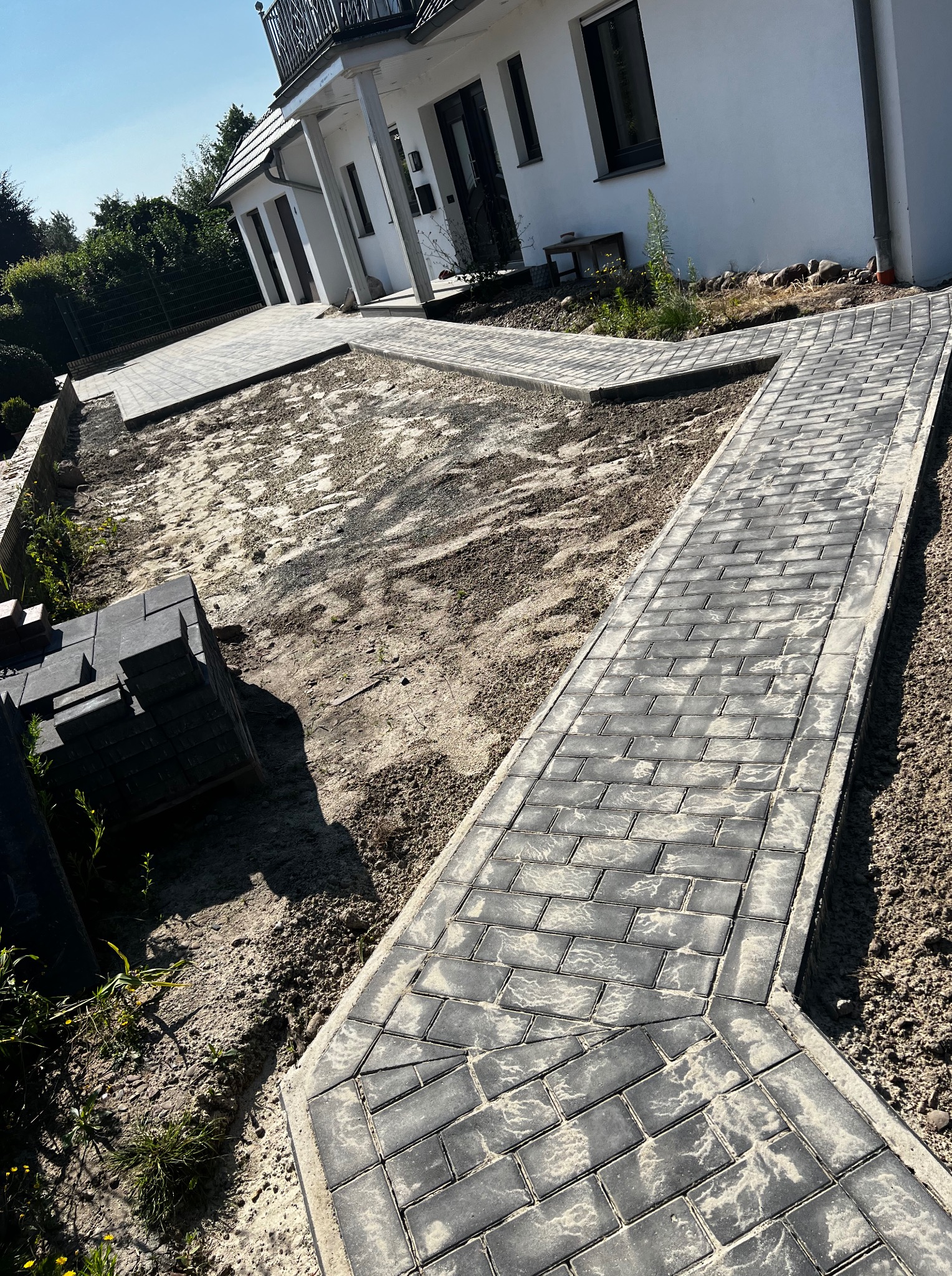 Układanie szarej kostki brukowej na ścieżce prowadzącej do białego domu. Stos kostki obok, ziemia przygotowana pod dalsze prace. Słoneczny dzień.