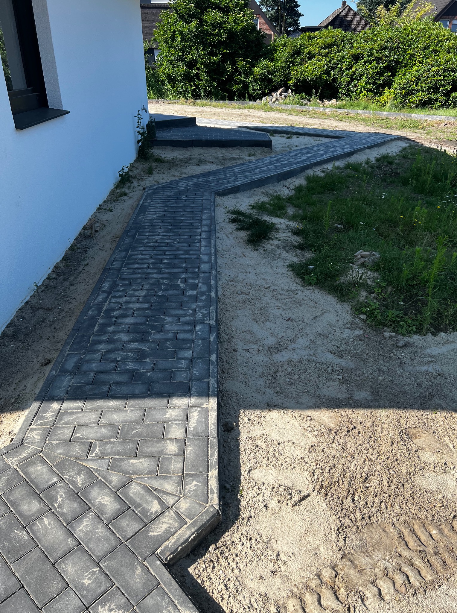 Ułożony chodnik z szarej kostki brukowej prowadzący wzdłuż białego budynku, otoczony piaskiem i zielenią. Widoczne ślady opon na piasku.