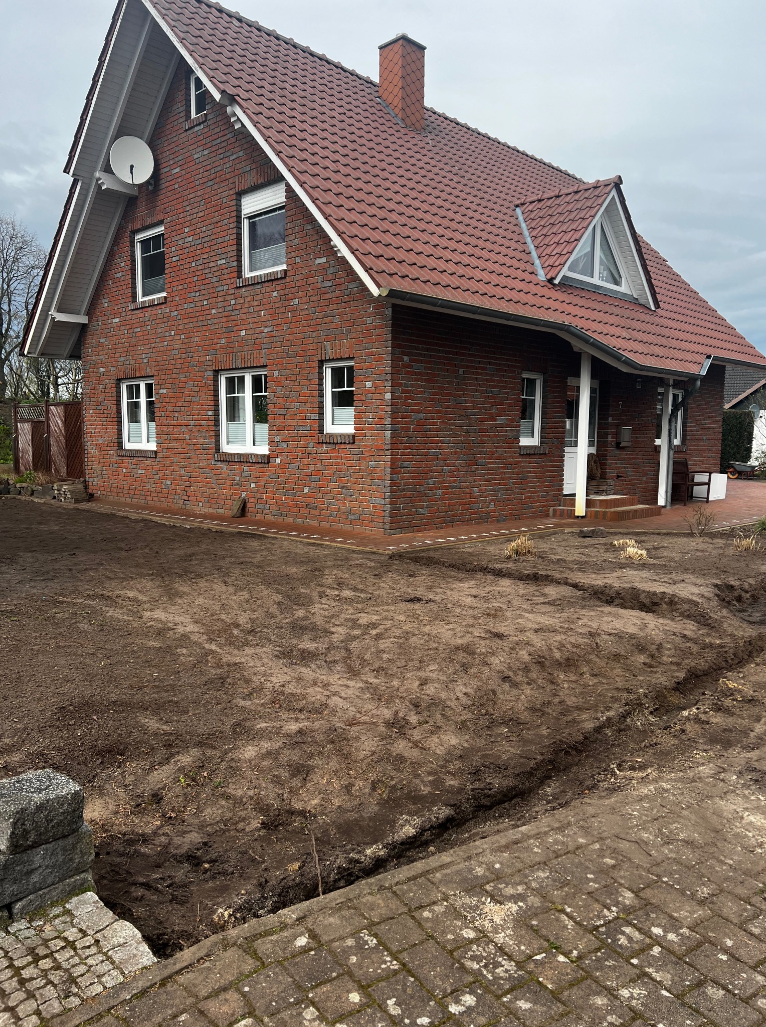 Dom z czerwonej cegły przed ogrodem w trakcie zakładania. Widoczne wykopane rowy pod instalacje. Na dachu antena satelitarna.