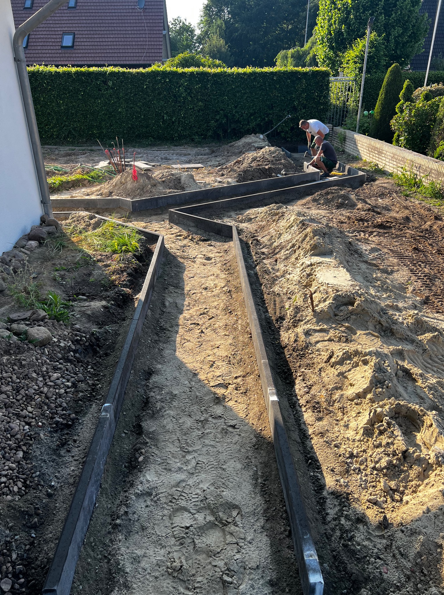 Prace ziemne przy zakładaniu ogrodu: dwóch mężczyzn montuje obrzeża rabat w zygzakowatym układzie. Widoczne narzędzia i przygotowany teren pod nasadzenia.