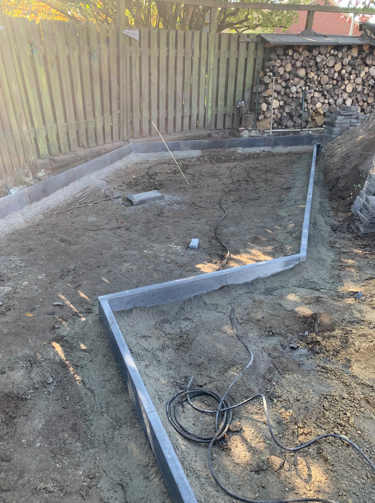 Przygotowanie podłoża pod ścieżkę z obrzeżami w ogrodzie. Widoczne ułożone krawężniki i ziemia przygotowana pod dalsze prace. W tle drewniany płot i ułożone drewno.