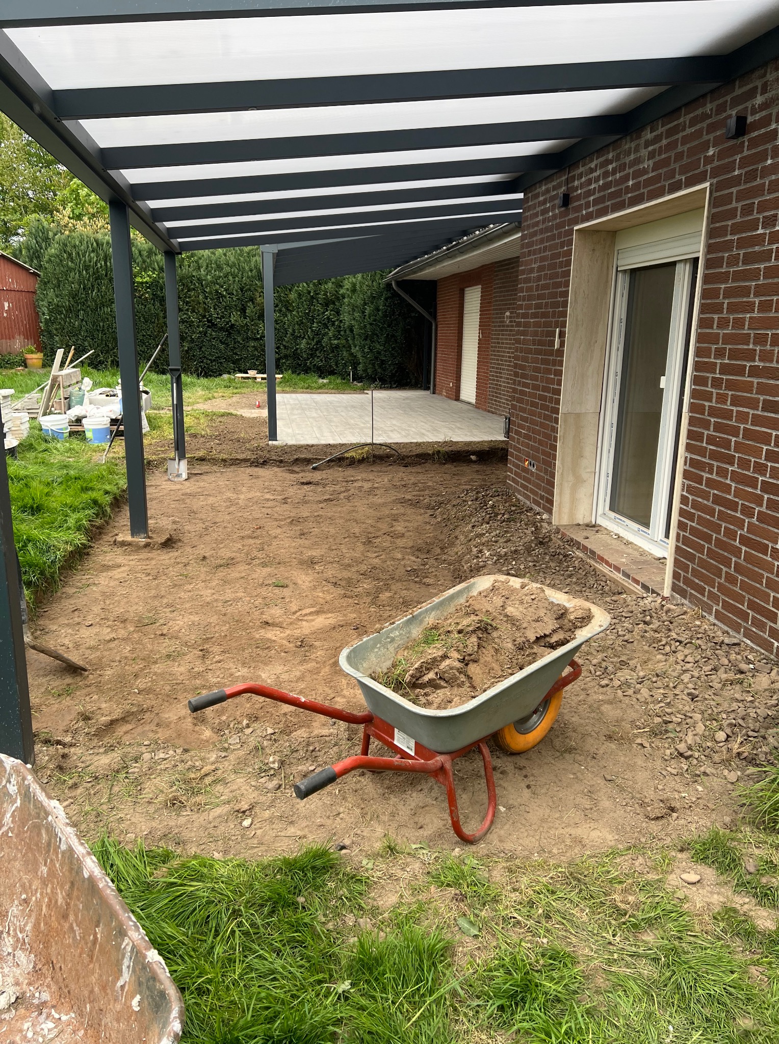 Przygotowanie terenu pod taras: taczka z ziemią, widoczne fundamenty pergoli i fragment ułożonej kostki. Etap prac ziemnych w ogrodzie.