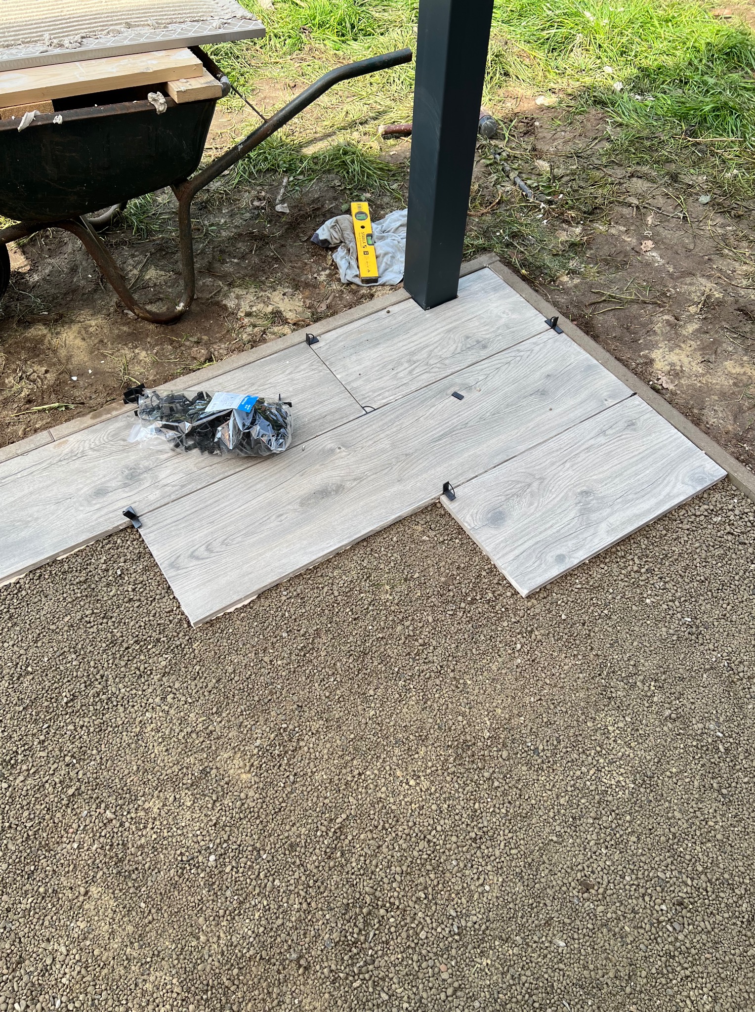 Układanie jasnych płytek imitujących drewno na żwirze, z użyciem poziomicy i wózka z materiałami. Etap prac w ogrodzie, widok z góry.